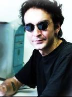 man  wearing dark sunglasses and a black t-shirt with a tatoo visible, sitting beside a computer with an impatient expression